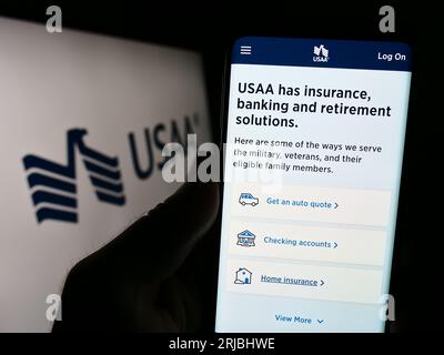 Person holding cellphone with webpage of United Services Automobile Association (USAA) on screen in front of logo. Focus on center of phone display. Stock Photo