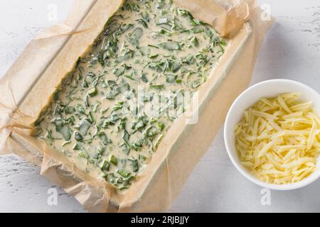 Spinat-Sauerampfer-Teig in einer Backform mit Pergament, eine Schüssel geriebenen Käse auf hellblauem Hintergrund, Draufsicht. Schritt für Schritt kochen. Stockfoto