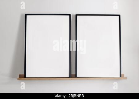 Vorlage für künstlerische Mockups. Zwei schwarze vertikale Bilderrahmen auf Bambus-Holzregal. Weißer Wandhintergrund. Poster zu Hause, minimalistisch skandinavisch Stockfoto