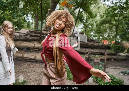 Junge und lächelnde afroamerikanische Frau im Boho-Outfit, die auf der Wiese im Retreat-Center tanzt Stockfoto