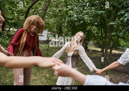 Glückliche multiethnische Freundinnen in Boho-Kleidung, die Hände halten und im Retreat Center tanzen Stockfoto