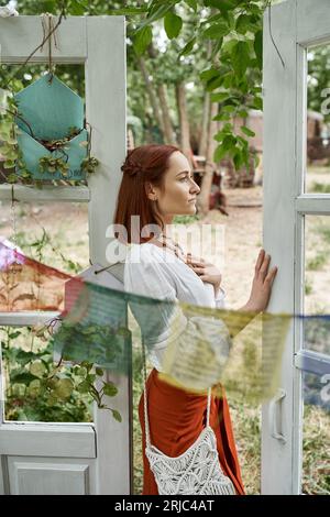 Schöne Rothaarige Frau in Boho-Outfit, die in der Nähe der Tür draußen im Retreat Center steht Stockfoto