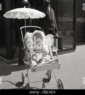 1960er Jahre, historisch, Sommer in der Stadt Draußen auf einem Bürgersteig, ein kleines Mädchen mit einer Motorhaube auf dem Kopf, sitzend in einem leichten Klappwagen der Ära, mit Schatten, London, England, Großbritannien. In dieser Zeit wurde der klappbare, leichte Kinderwagen – oder Buggy, wie er später auch genannt wurde – populär und übernahm die Rolle des größeren Kinderwagens, des traditionellen großen Kinderwagens mit Rädern. Stockfoto