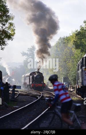 Ein Radfahrer auf einem ausgewiesenen Radweg, der bei Bitton die Avon Valley Railway überquert, während sich eine Dampfeisenbahn nähert. Stockfoto