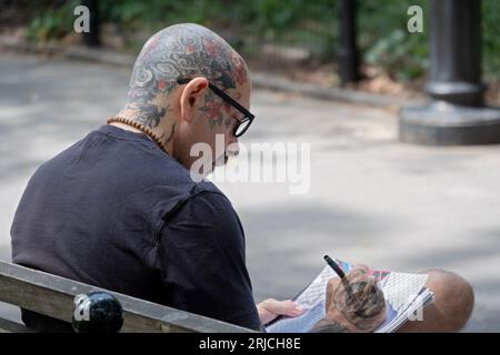 Ein Mann mittleren Alters mit einem vollständig tätowierten, rasierten Kopf zieht, während er auf einer Bank im Washington Square Park in Greenwich Village sitzt. Stockfoto
