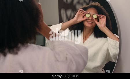 Glückliche afroamerikanische Frau, lustiges Mädchen im Bademantel, das Spaß hat, um Spiegelreflexionen herumzuspielen, frische Gurkenschnitte, Augen, die lächeln Stockfoto