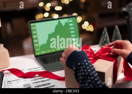 Eine Analytikerin an ihrem Schreibtisch arbeitet an einem Laptop, der Statistiken und Diagramme während der Weihnachtszeit anzeigt. Sie arbeitet an der Tischverpackung, mit der sie ein Geschenk macht Stockfoto