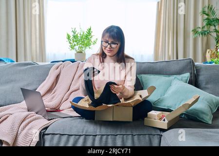 Reife Frau, die zu Hause sitzt, Pappkarton auspackt und online einkauft Stockfoto