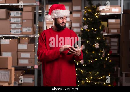 Männlicher Lagerarbeiter, der zu weihnachten mit einem digitalen Tablet im Lagerhaus arbeitet. Weihnachtslieferung Stockfoto