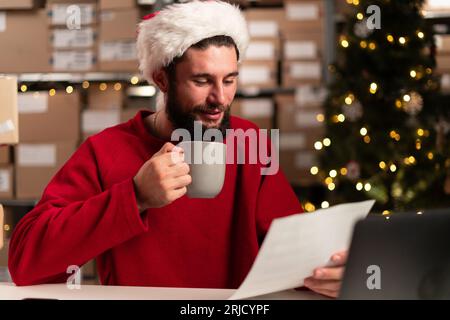 Weihnachtsgeschäft. Happy Modern Wear Santa Hut mit Laptop und Paketen mit Dokumenten und Kaffee im Lager. Stockfoto