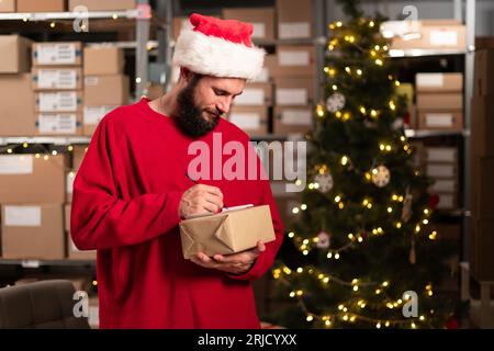 Glücklicher Weihnachtsmann Verpackung Geschenkkarton Schreibadresse Vorbereitung des Postversandpakets im Werkstattlager. Frohe Weihnachten Versand Lieferung Stockfoto