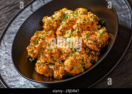 Bang Bang Brokkolibisse, knusprige gebackene Brokkoliröschen mit Mayo-Sriracha-Sauce in schwarzer Schüssel auf dunklem Holztisch, Nahaufnahme Stockfoto