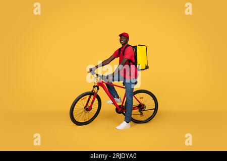 Fröhlicher, attraktiver, tausendjähriger afroamerikanischer Boy-Kurier in Uniform mit Fahrradtouren im Rucksack Stockfoto