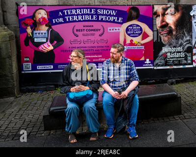 Eine Szene in Edinburghs Royal Mile während der letzten Woche des Edinburgh Festival Fringe 2023. Ein Paar sitzt auf einer Bank neben Festivalplakaten. Stockfoto