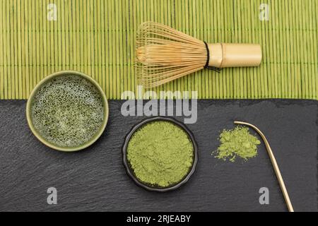 Bambusquirl auf grüner Serviette aus Holzstäben. Matcha-Teepulver und Matcha-Tee auf Steinbrett. Flach liegend. Stockfoto