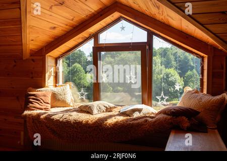 Ein Fenster mit einer breiten gemütlichen Fensterbank im zweiten Stock eines Holzhauses. Die Fensterbank ist mit einer Pelzdecke und Kissen bedeckt. Die Fensteröffnung Stockfoto