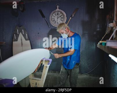 Seitenansicht eines erwachsenen blonden Hairmaster in einer Atemschutzmaske, die weißes Surfbrett in Formgestelle während der Arbeit in der Werkstatt mahlt Stockfoto