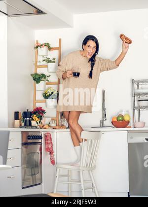 Ein voller Körper von Frauen in Heimkleidung, die auf einem Stuhl in der Nähe der Theke stehen und Kaffee trinken, während sie am Wochenende zu Hause den Morgen genießen Stockfoto