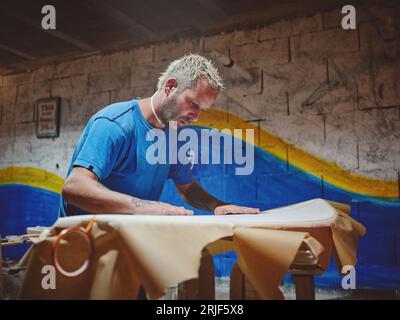 Seitenansicht eines bärtigen blonden männlichen Masters, der handgefertigtes Surfbrett in Stoff wickelt, während er das Brett in der Werkstatt verzierte Stockfoto