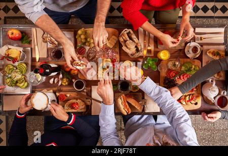 Von oben sehen Sie eine Gruppe von gesichtslosen Freunden, die köstliche Snacks essen, während sie sich am Holztisch mit Getränken in der hellen Küche treffen Stockfoto