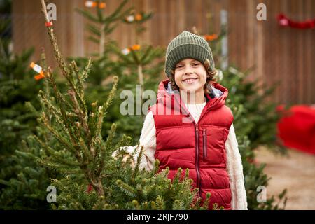 Fröhlicher kleiner Junge in stylischer Oberbekleidung und Hut lächelnd und mit Blick auf die Kamera, während er am Wintertag in der Nähe eines Nadelbaums auf dem Markt steht Stockfoto