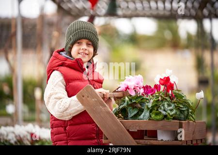 Niedlicher Junge in Strickmütze, der in der Nähe eines Holzwagens steht, mit lebhaften blühenden Blumen in Töpfen im botanischen Garten Stockfoto