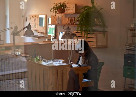Durch ein Glas seriöser junger Handwerkerin mit dunklen Haaren in lässiger Kleidung, die auf einer Holzbank sitzt, während sie Schmuck mit verschiedenen Handbüchern kreiert Stockfoto