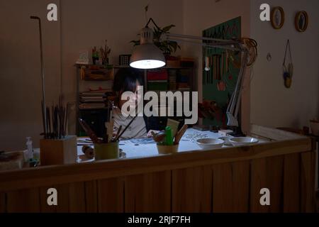 Weiblicher Meister, der am Tisch im Schmuckstudio sitzt und von der Lampe beleuchtet wird und während der Arbeit nach unten schaut Stockfoto