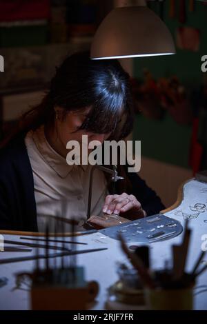 Von oben eine fokussierte erwachsene ethnische Goldschmiede, die auf der Werkbank sitzt und in der traditionellen Werkstatt mit Säge goldenen Schmuck repariert Stockfoto