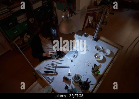 Hoher Winkel aus anonym plattiertem Metall mit Handfeile am Schreibtisch mit Lampe und verschiedenen Werkzeugen in dunkler Werkstatt Stockfoto