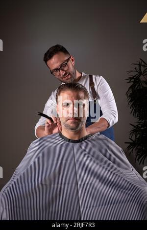 Professioneller männlicher Barbier mit Rasiermesserhals eines ernsten Mannes, der im Stuhl auf grauem Hintergrund im hellen modernen Friseursalon sitzt Stockfoto