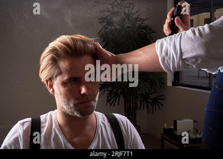 Ausgereifter gutaussehender, selbstbewusster Mann, der wegschaut, und nicht erkennbarer Friseur, der mit Haaren des Kunden mit Spray arbeitet Stockfoto