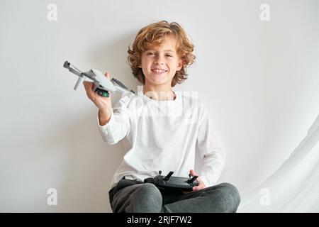 Zufriedenes blondes Kind mit Spielzeugdrohne in erhobener Hand, das die Kamera gegen die weiße Wand schaut Stockfoto