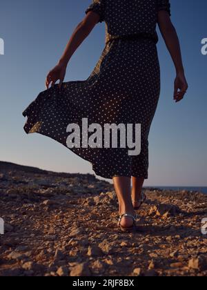 Von unten eine nicht erkennbare Frau in stilvollem Polka-Dot-Kleid, die bei Sonnenuntergang an felsigem Ufer in der Nähe des Meeres spaziert Stockfoto