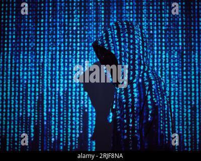 Seitenansicht eines anonymen Hackers im Hoodie, der in blauer Projektion des Programmiercodes in einem dunklen Lichtraum steht Stockfoto