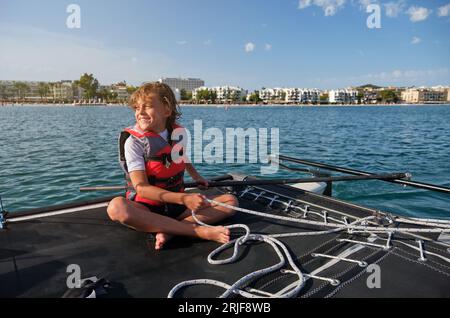 Voller Inhalt Barfuß-Kleinkind mit blondem Haar in der Schwimmweste lächelnd und wegschauend, während es auf dem Boot sitzt und im Sommer an einem Seil zieht Stockfoto