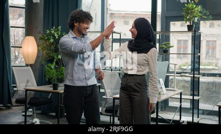 Zwei multirassische Kollegen glückliche Firmenkollegen arabischer indischer Mann Geschäftsmann, der hohe fünf muslimische Geschäftsfrau in Hijab Geschäftsleuten gibt Stockfoto
