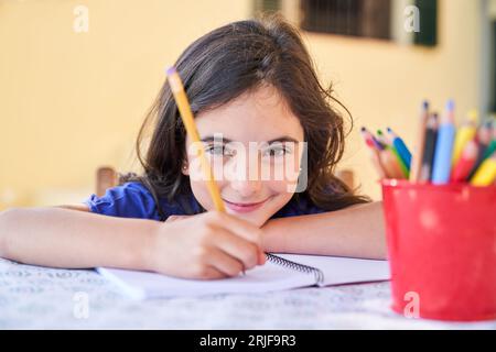 Kreatives, süßes Schulmädchen, das Notizen auf Papier mit Bleistift im Copybook macht, während es Zeit zu Hause verbringt und die Kamera betrachtet Stockfoto