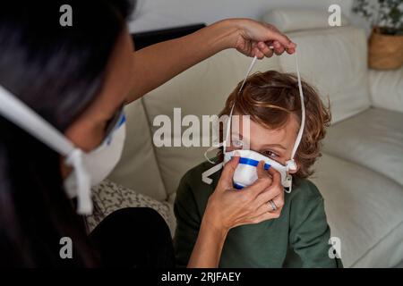 Hohe Winkel der Ernte anonyme Mutter mit dunklen Haaren, die medizinische Maske auf das Gesicht des kranken Sohnes sitzend auf dem Sofa während der Coronavirus-Pandemie Stockfoto