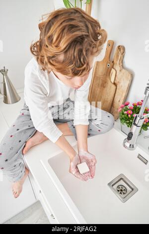 Ein hochwinkeliger Körper eines Barfußjungen im Pyjama, der auf der Theke in der Nähe von Schneidebrettern sitzt und morgens in der Küche die Hände mit Seife wäscht Stockfoto