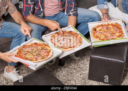 Von oben aus zeigen anonyme männliche Freunde in lässiger Kleidung, die im Wohnzimmer sitzen und auf verschiedene Arten von Pizza zeigen Stockfoto