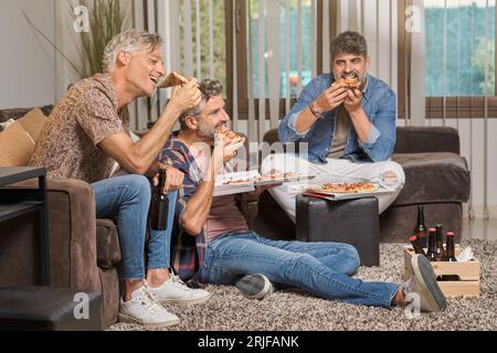 Gruppe reifer männlicher Freunde, die leckere Pizza essen und Bier trinken, während sie zu Hause im Wohnzimmer sitzen Stockfoto