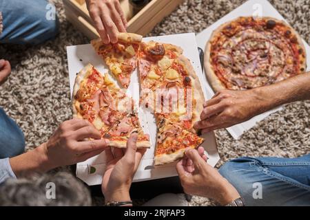 Von oben gesichtslose männliche Freunde, die sich treffen und köstliche Pizzascheiben nehmen, während sie gemeinsam am Wochenende feiern Stockfoto