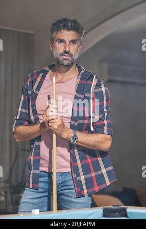 Nachdenklicher, reifer bärtiger Mann in zwanglosem Outfit mit Armbanduhr, die in der Nähe des Billardtisches steht, mit einem Hinweis, während sie in einem rauchigen Apartment wegschaut Stockfoto