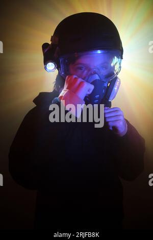 Ernsthaftes Kind in Schutzgasmaske mit leuchtender Taschenlampe, das auf die Kamera mit geballten Fäusten schaut, die bereit für den Kampf sind, während es dunkle Minze gegen Farbe ausraubt Stockfoto