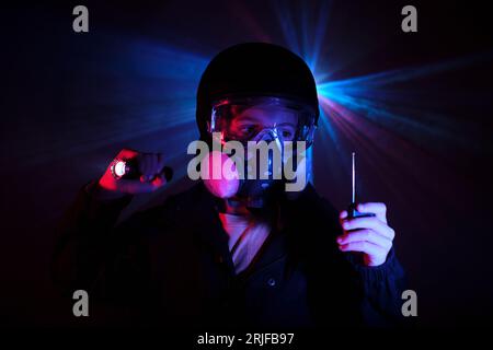 Schläger in Atemschutzmaske mit tragbarem Walkie-Talkie in der Hand leuchtende Taschenlampe, während dunkle Minze mit roten und blauen Neonlichtern beraubt wird Stockfoto