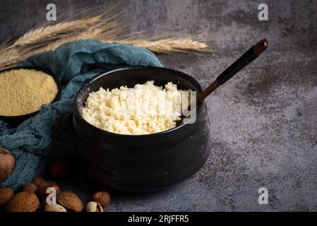 Roher und gekochter Couscous in einer schwarzen Keramikschale auf einem rustikalen grauen Hintergrund, Ansicht von der Tischplatte Stockfoto