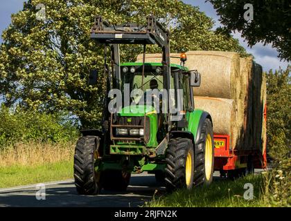 Woburn, Beds.UK - 19. August 2023: Erntezeit. John Deere Traktor, der einen Anhänger mit großen Rundballen zieht, fährt mit einem Auto auf einem englischen Land Stockfoto