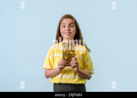 Ein fröhliches Mädchen im Teenageralter hält ein Geschenk in einem vergoldeten Paket mit einer Schleife in den Händen. Hellblauer Hintergrund Stockfoto