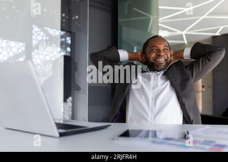 Ein gut gefertigter Geschäftsmann hat die Arbeit erfolgreich abgeschlossen, ruht träumerisch und nachdenklich im Büro, der Mann wirft seine Hände hinter den Kopf und visualisiert die zukünftigen Siege und Erfolge. Stockfoto